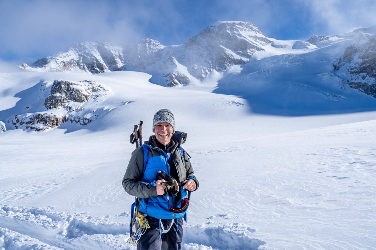 Person mit Kamera auf einem Gletscher, dahinter verschneite Gipfel