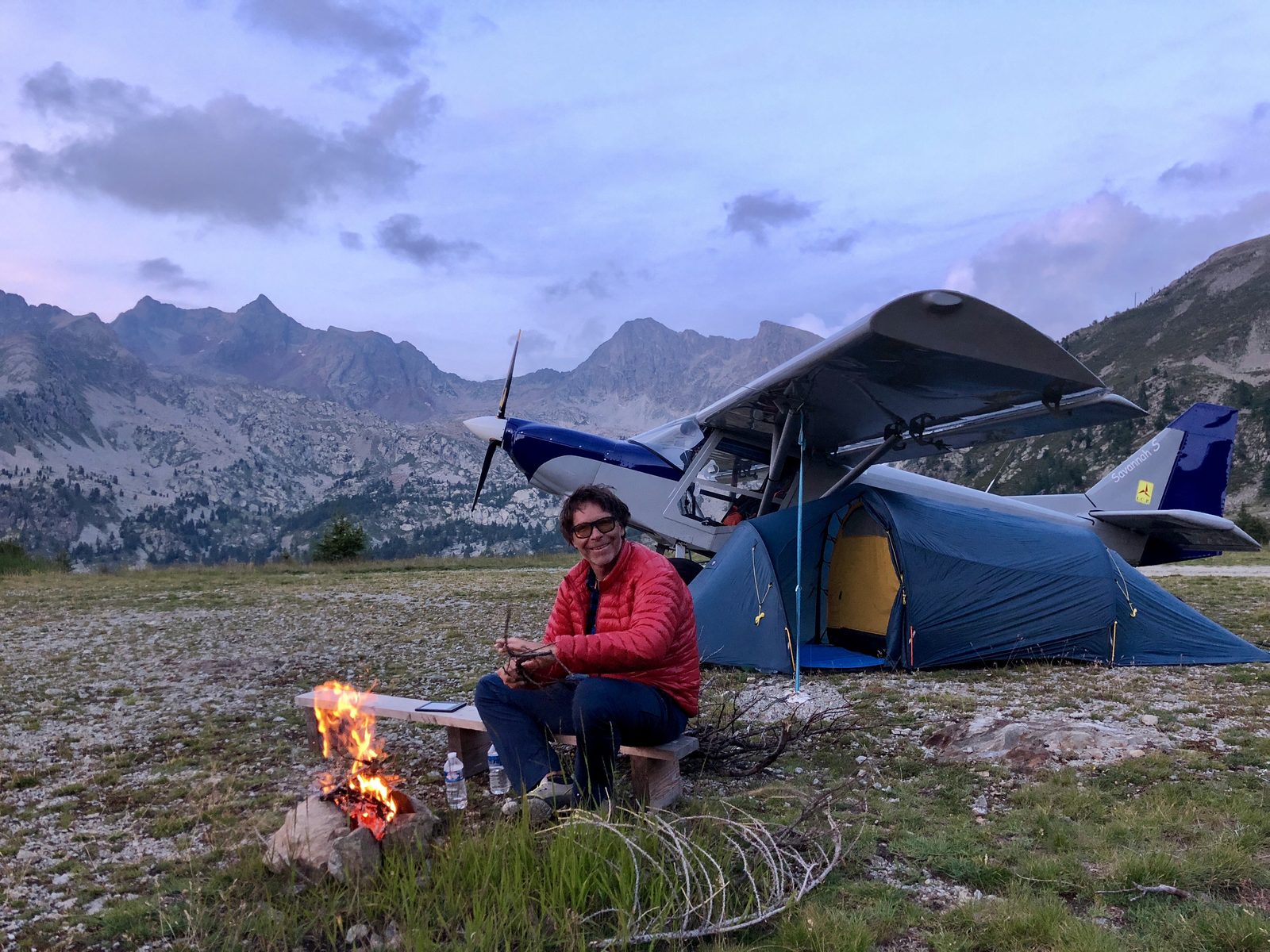 Mann in roter Jacke am Lagerfeuer, dahinter ein Zelt und ein Leichtflugzeug vor Berggipfeln in der Dämmerung