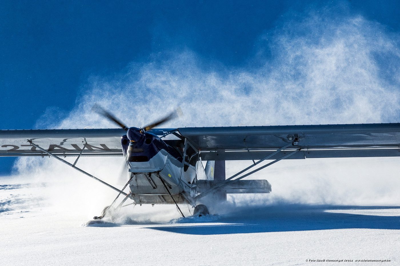 Leichtflugzeug landet auf einer verschneiten Fläche, dahinter Berge