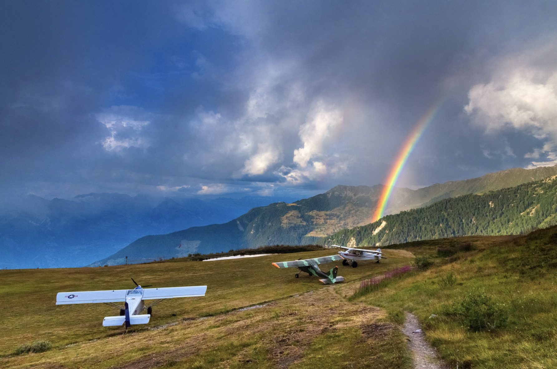 Drei Leichtflugzeuge auf einer Bergwiese, darüber ein Regenbogen
