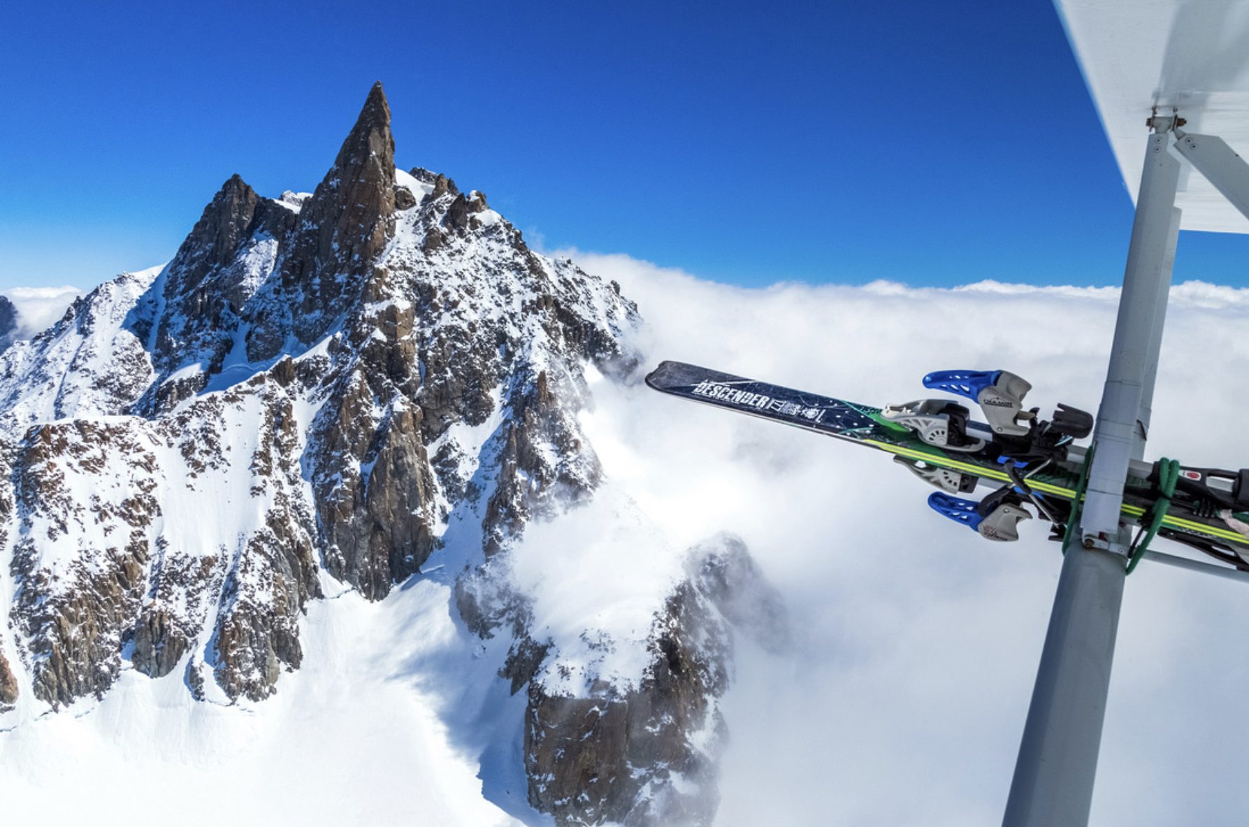 Blick aus dem Flugzeug: Ski an der Flügelstrebe, daneben ein Felsgipfel über dem Nebel