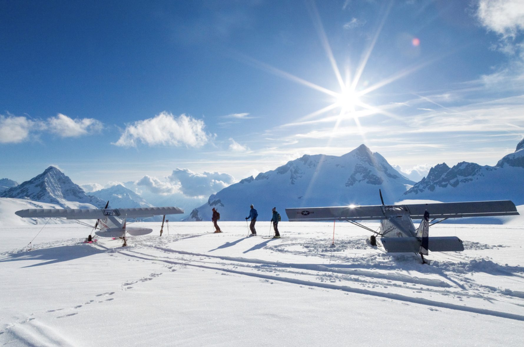 Zwei Leichtflugzeuge auf einem Gletscher, dazwischen drei Personen mit Tourenski, Gegenlicht