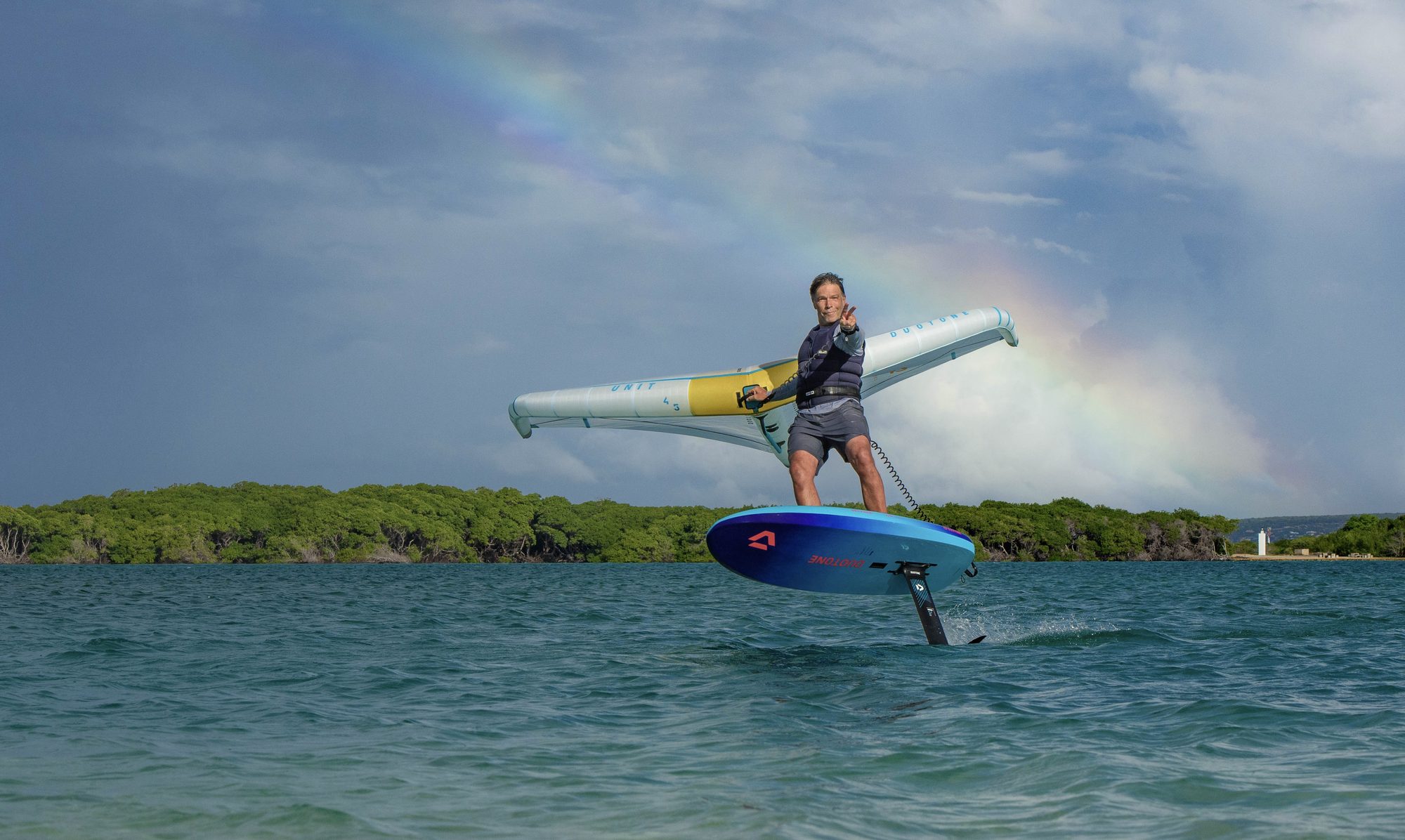 Roland beim Wingfoilen vor einem Regenbogen, er zeigt das Peace-Zeichen