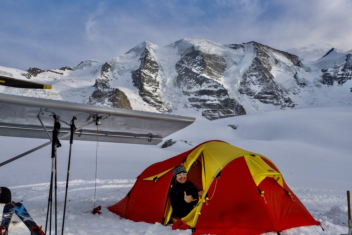 Rot-gelbes Zelt im Schnee unter dem Flügel eines Leichtflugzeugs, dahinter vergletscherte Berge
